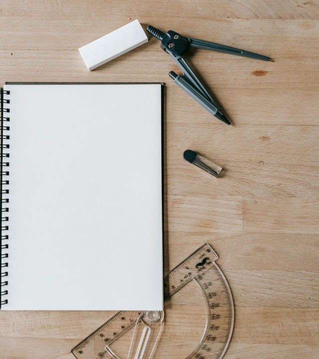 Flat lay of school supplies including notebook, compass, eraser, and protractor on wooden desk.